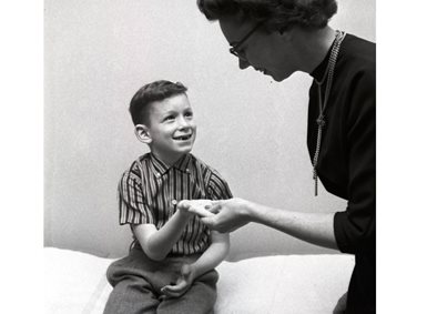 En liten gutt med tannløst smil holder frem hånden med en melketann i. En voksen dame kikker ned på den. Bildet er i sort-hvitt.